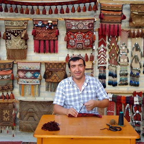 A man sits at a table, weaving, with colorful textiles and wall hangings displayed behind him.