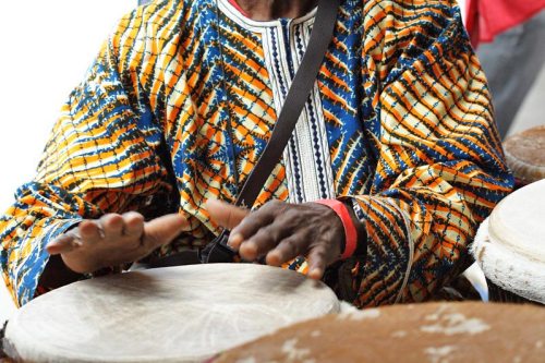 A person in a colorful patterned shirt plays a drum, hands poised above the instrument.