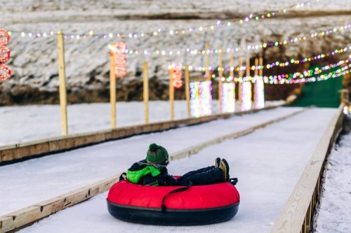 A child in a red snow tube slides down a snowy slope, surrounded by colorful holiday lights.