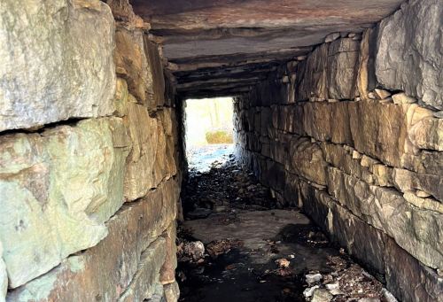 A stone tunnel with rough walls, leading to a bright opening at the far end, surrounded by debris and natural light.