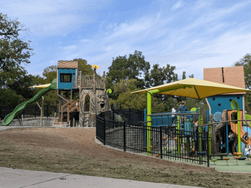 A colorful playground with slides, climbing structures, and shaded areas, surrounded by grass and trees under a blue sky.