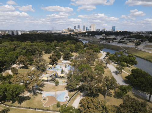 Aerial view of a park with playgrounds and trees, overlooking a city skyline and a river in the background.