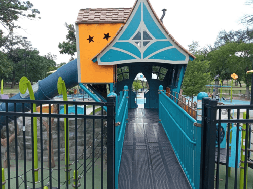 Colorful playground structure with a slide, surrounded by a fence and greenery in the background.