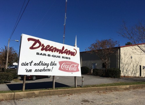 Sign for Dreamland Bar-B-Que Ribs with a Coca-Cola logo, set against a clear blue sky and a building in the background.