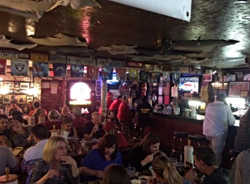 A lively bar scene with patrons dining and socializing, surrounded by memorabilia and a TV showing a sports game.