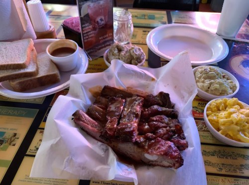 A table set with a plate of ribs, sides of macaroni and cheese, coleslaw, bread, and a drink in a casual dining setting.