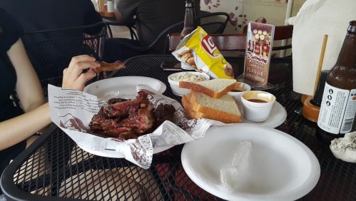 A table with a plate of ribs, bread, coleslaw, chips, and a drink, with a hand reaching for a rib.