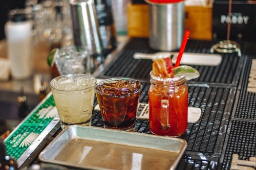 Three cocktails on a bar: a margarita, a dark drink in a glass, and a colorful drink with a garnish and straw.