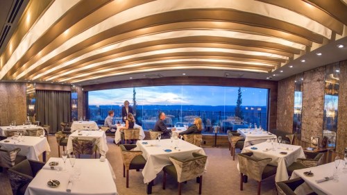 Elegant restaurant interior with tables set for dining, large windows showcasing a scenic view at sunset.