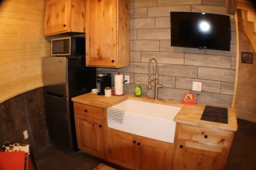 A cozy kitchen with wooden cabinets, a farmhouse sink, and modern appliances, including a TV mounted on the wall.