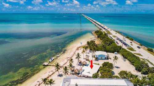 Aerial view of a beach with palm trees, a pier, and a road, featuring a highlighted building near the shore.