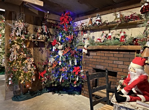 A cozy room decorated for Christmas, featuring two festive trees, ornaments, and a Santa figure by a brick fireplace.