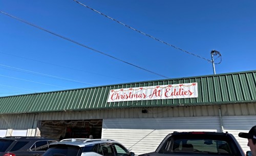 Sign on a building reads "Christmas At Eddie's" against a clear blue sky, with parked cars in the foreground.