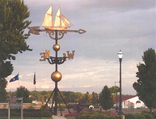 A decorative weather vane featuring a ship and the word "Welcome," set against a cloudy sky and green trees.