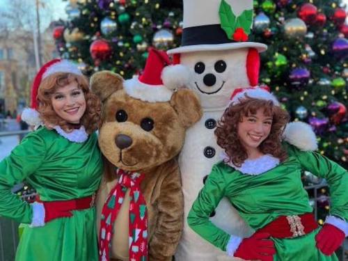 Two women in green dresses and Santa hats pose with a large teddy bear and a snowman in front of a decorated Christmas tree.