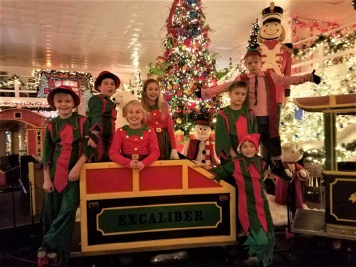 A group of children dressed as elves poses in front of a decorated Christmas tree and festive decorations.