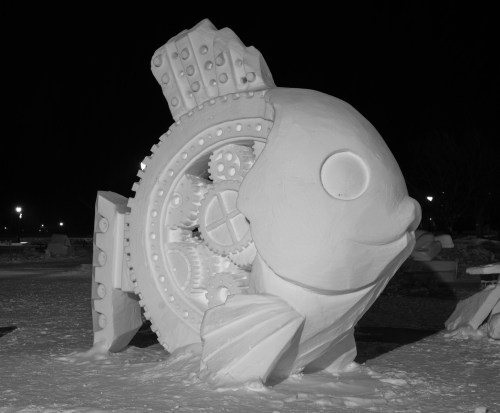 A large, intricately carved snow sculpture of a fish with mechanical gears, set against a dark night background.