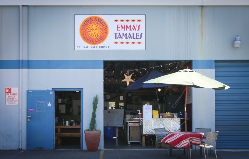 Exterior of Emma's Tamales, featuring a colorful sign, outdoor seating, and a blue door under a sunny sky.
