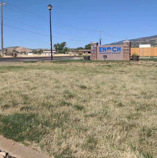 Sign for Enoch City in a grassy area, with a lamp post and distant hills under a clear blue sky.