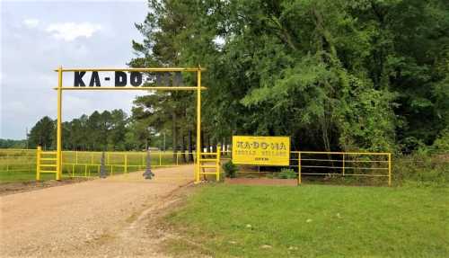 A yellow entrance gate with "KA-DO-MA" sign, surrounded by trees and a gravel path leading into a rural area.