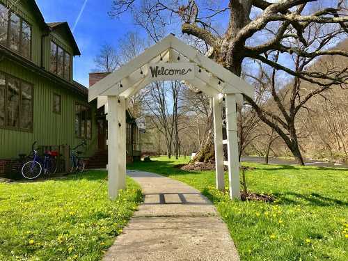 A white archway with "Welcome" sign leads to a green house, surrounded by trees and a grassy path.