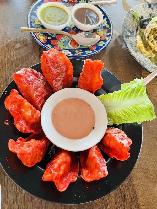 A plate of vibrant red dumplings arranged around a small bowl of dipping sauce, garnished with lettuce.