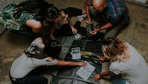 A group of five people examining evidence on a black tarp in a dimly lit room, focused on various documents and tools.