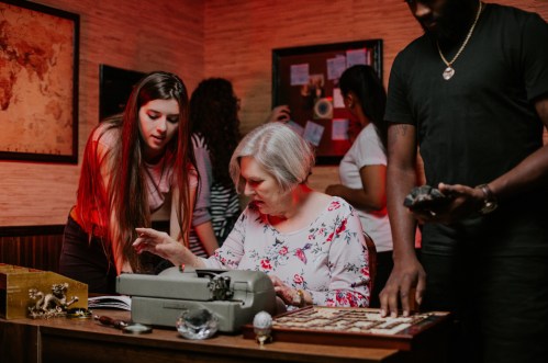 A group of people collaborates in a dimly lit room, examining clues and a typewriter during an escape room challenge.