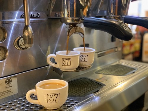 Two white coffee cups with "S7" printed on them are positioned under an espresso machine, filling with coffee.