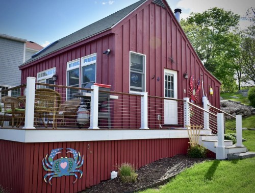 A red cottage with a deck, featuring a crab decoration and outdoor seating, surrounded by green grass and trees.