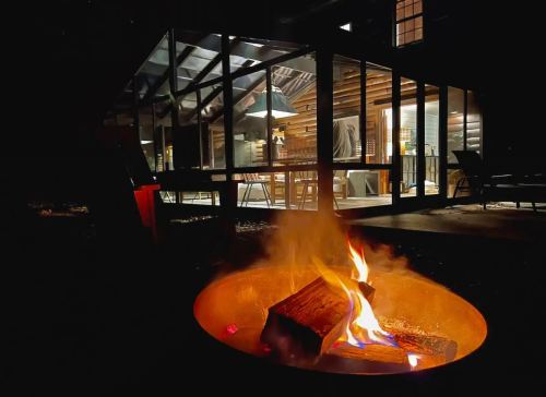 A cozy outdoor fire pit with flames, glowing warmly in front of a glass-enclosed patio at night.