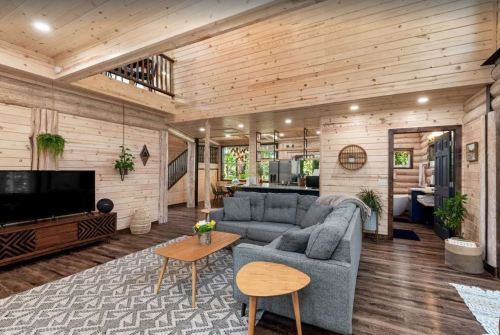 Cozy living room with wooden walls, a gray sofa, coffee table, and a TV, leading to a kitchen and staircase.