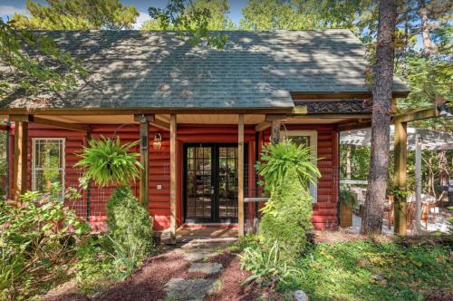 A charming red cabin surrounded by greenery, featuring a welcoming entrance with potted ferns and stone pathway.