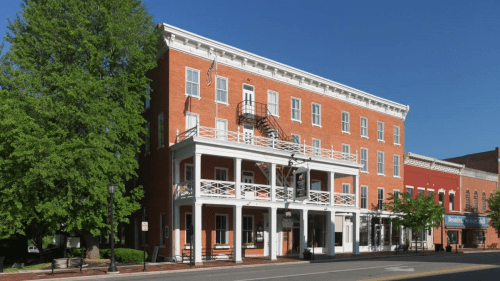 Historic red brick building with a white balcony and fire escape, surrounded by trees and blue sky.