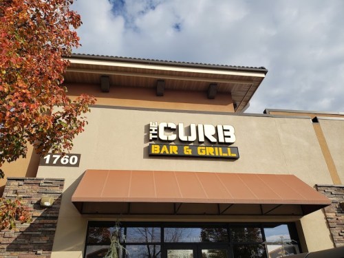 Sign for "The Curb Bar & Grill" on a building with a cloudy sky and autumn leaves in the foreground.