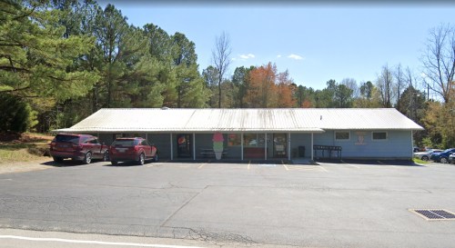 A single-story building with a metal roof, surrounded by trees and parked cars, on a clear day.