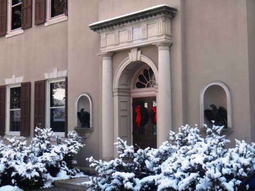A snowy entrance of a house with a decorative archway, red wreaths, and evergreen bushes.