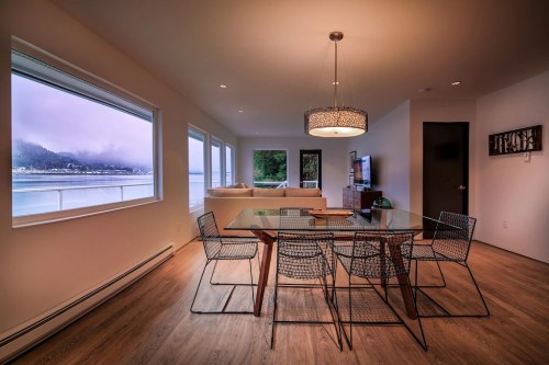 Modern dining area with a glass table, wire chairs, and large windows overlooking a scenic view of water and mountains.