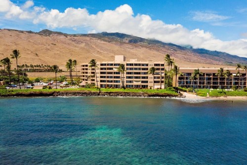 Aerial view of a beachfront hotel with palm trees, mountains, and clear blue water under a partly cloudy sky.
