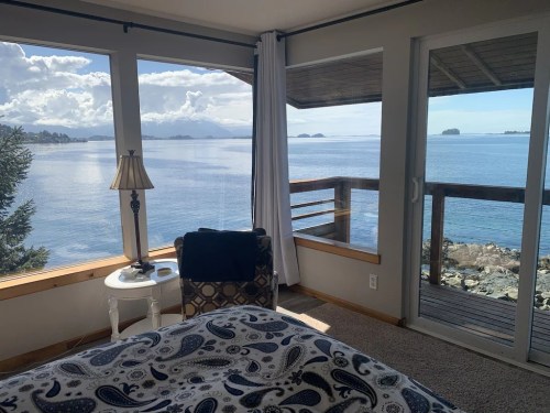 Cozy bedroom with a view of the ocean and islands, featuring a chair, lamp, and large windows letting in natural light.