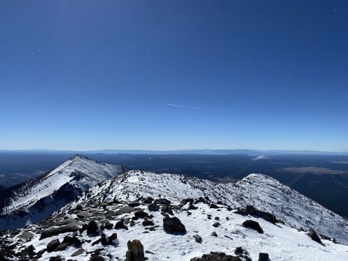 Snow-covered mountain peaks under a clear blue sky, with distant valleys and a faint contrail in the horizon.