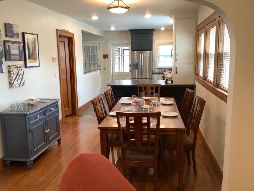 A cozy dining area with a wooden table set for a meal, leading to a modern kitchen in a well-lit home.
