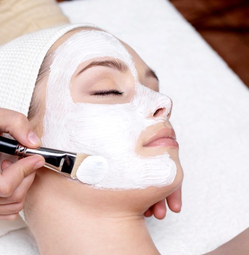 A person receiving a facial treatment, with a white mask being applied to their face using a brush.