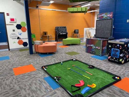 A colorful game room featuring a mini golf setup, seating areas, and interactive game stations on a carpeted floor.