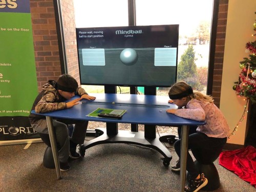 Two children with blindfolds lean over a table, focused on a screen displaying a game called "Mindball."