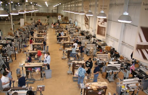A spacious workshop filled with people working at various woodworking stations, surrounded by tools and materials.