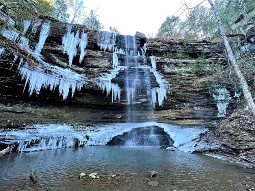 A frozen waterfall with icicles hanging from rocky cliffs, surrounded by trees and a calm pool of water below.