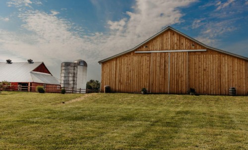 A scenic farm with a wooden barn, silo, and green grass under a blue sky with scattered clouds.