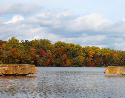 A serene lake surrounded by vibrant autumn foliage and cloudy skies, with stone structures in the foreground.