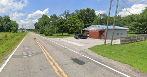 A rural road with a small building and a parked car, surrounded by trees and greenery under a blue sky.